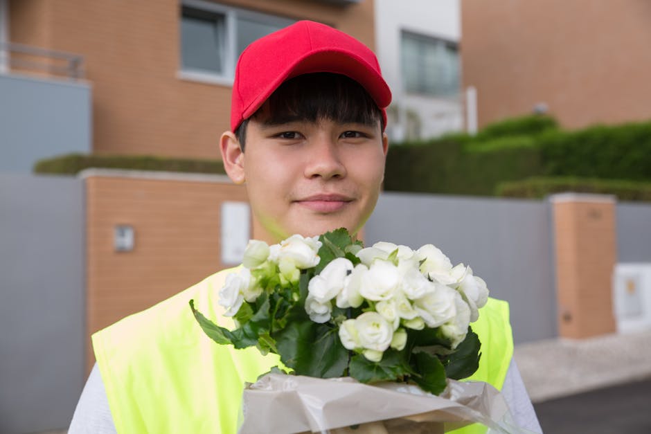 A young delivery person wearing a bright yellow high-visibility vest and a red cap is standing outdoors in front of modern residential buildings with brick and concrete exteriors. They are holding a b