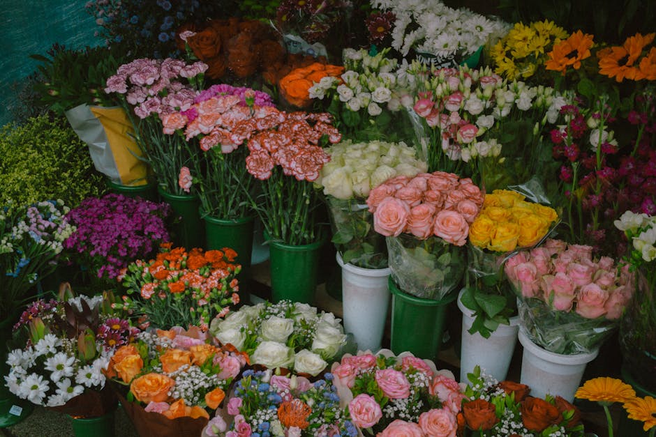 A vibrant display of fresh flowers arranged in various bouquets and buckets at a flower shop. The assortment includes pink, orange, white, yellow, and purple blossoms such as roses, carnations, daisie