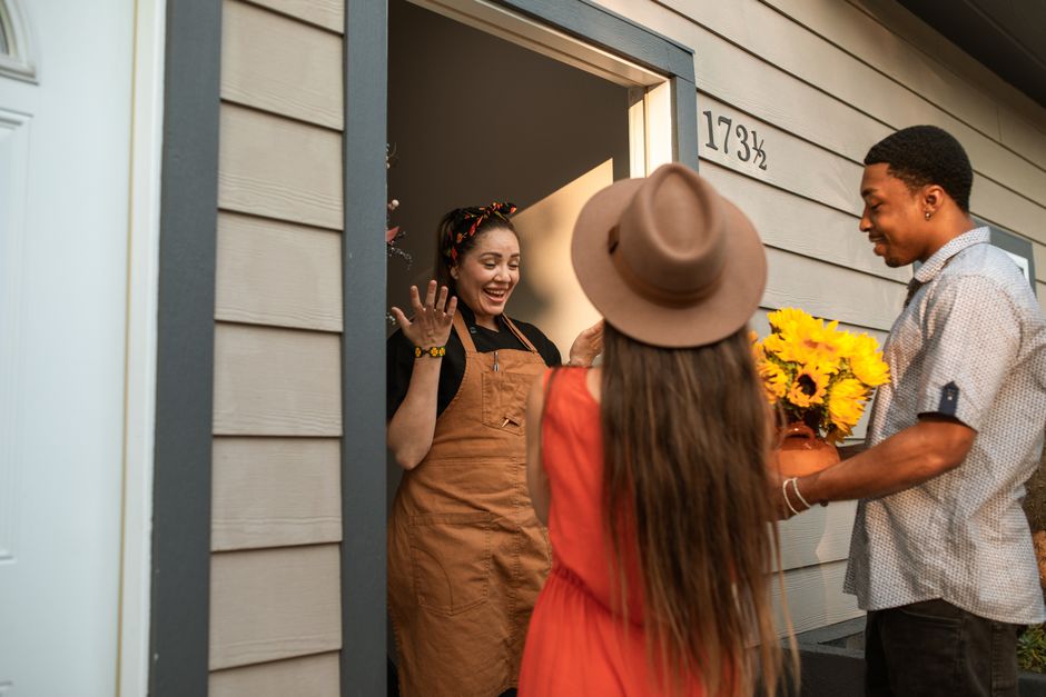 A woman in an orange apron, smiling warmly, stands at her front door greeting two friends who are holding a bouquet of vibrant yellow sunflowers. The woman at the doorstep has dark hair tied back with