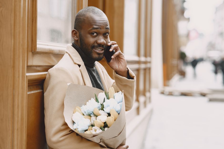 A man with a dark complexion and short hair is standing outdoors against a wooden panel wall, holding a bouquet of flowers wrapped in brown paper with pastel-colored blooms in shades of blue, cream, a